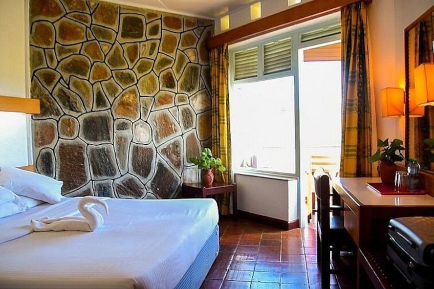 A hotel room with a stone wall, a bed with white linens, a desk with a lamp, and a window with curtains. 3