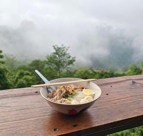 A bowl of ramen with chopsticks on a wooden table with a misty mountain backdrop. 3