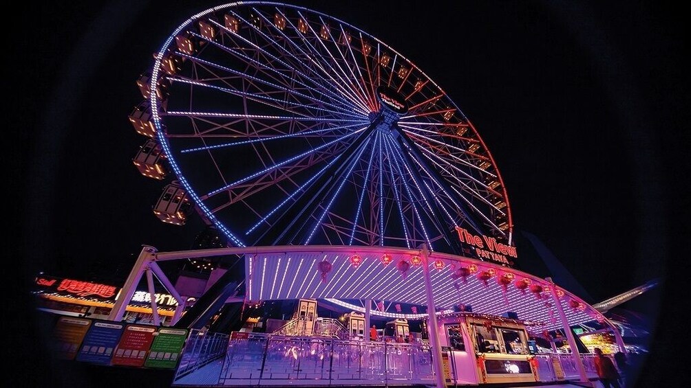 Una grande ruota panoramica di notte illuminata da luci colorate, con un'attrazione coperta sottostante. 1