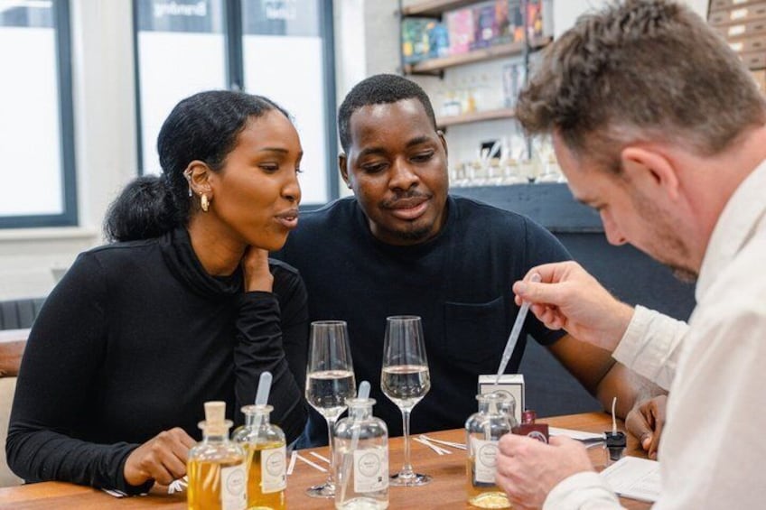 Three people seated at a table with wine glasses, bottles, and a tasting spoon. 2