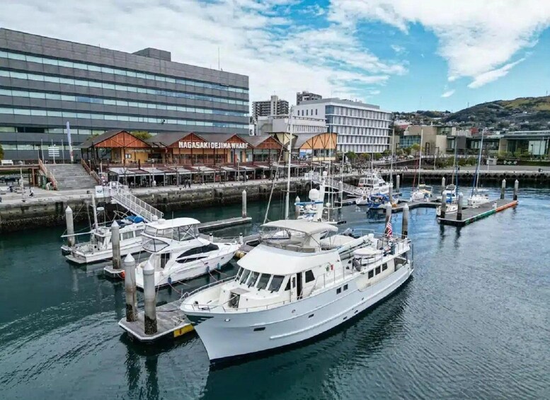 A marina with several boats docked, a building with a sign reading 'Nagasaki Dejima Wharf,' and a modern building in the background. 3