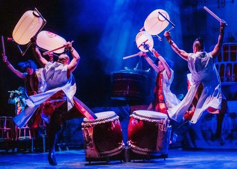 A group of performers with drums and tambourines, in a stage setting with blue lighting. 2
