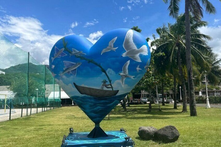 A heart-shaped sculpture with a boat and seagulls, set on grass with palm trees in the background. 4