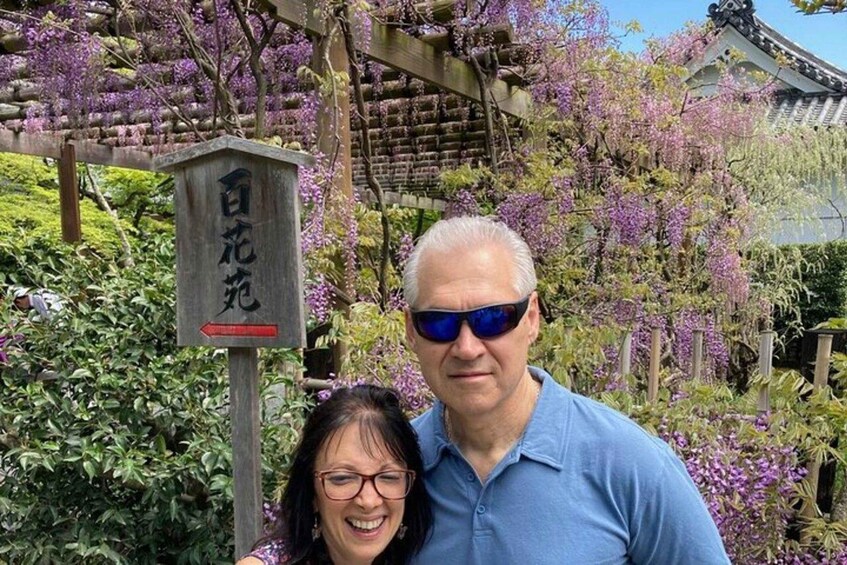 Una pareja de pie frente a un cartel con caracteres japoneses, rodeada de glicinias en flor. 3