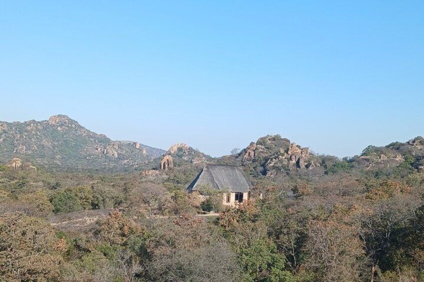 A house with a thatched roof surrounded by dry vegetation and rocky hills. 1
