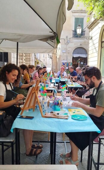 A group of people painting outdoors at a table with easels and paint supplies. 2