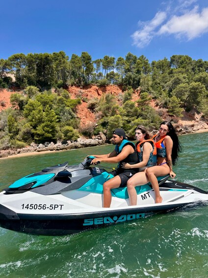 A jet ski with three people on it, moving on a body of water with a rocky shoreline and trees in the background. 2