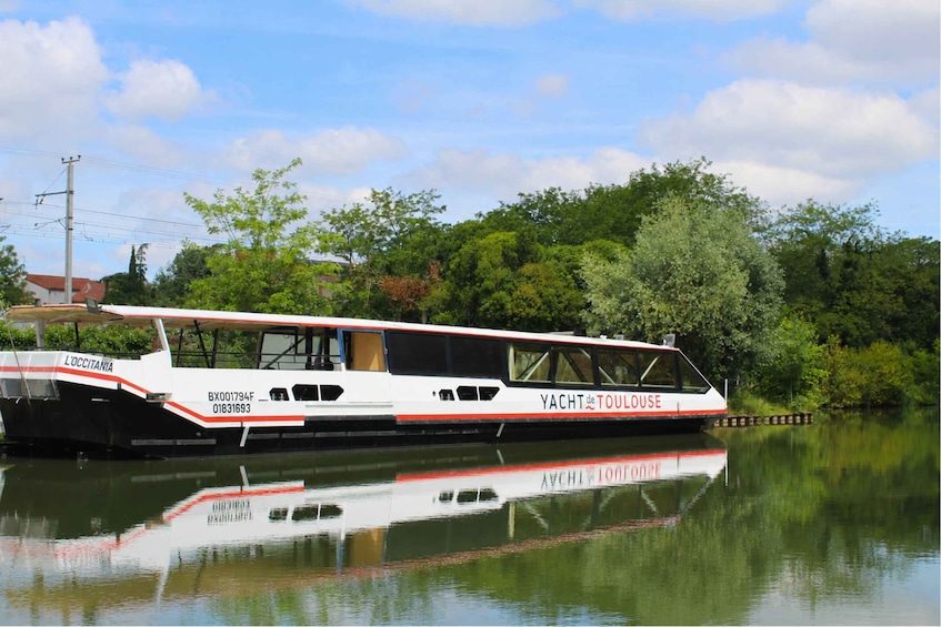 A white and red boat named 'YACHT de TOULOUSE' docked by a calm river, surrounded by lush greenery and a clear sky. 1