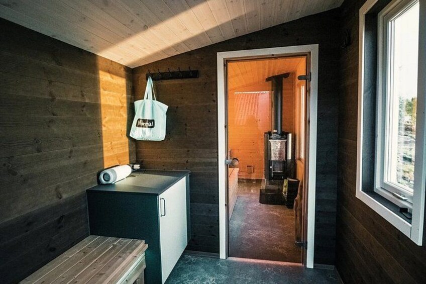 A wooden cabin interior with a small kitchenette, a wooden bench, a hanging bag, and a door leading to a room with a wood-burning stove. 4