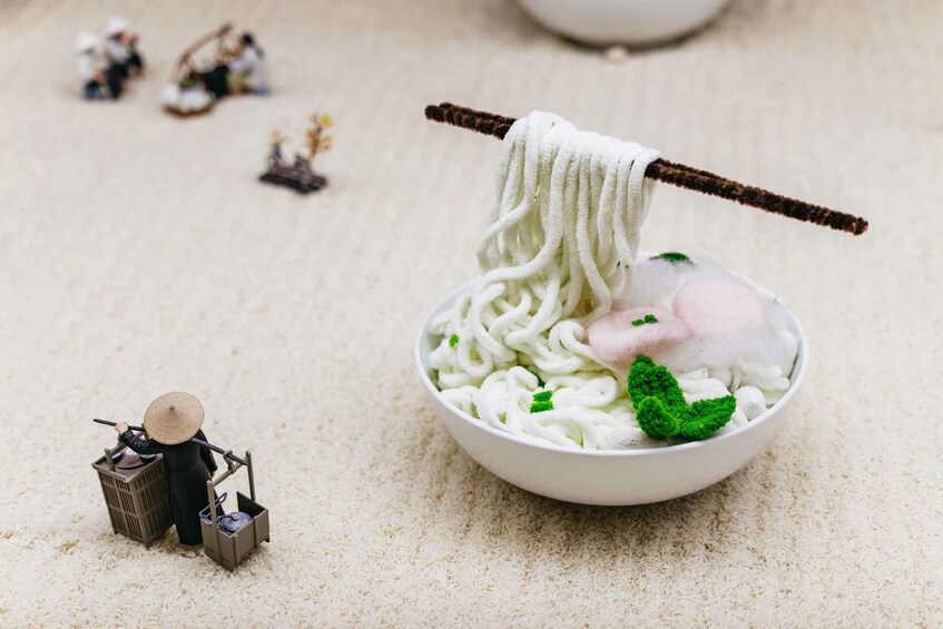 A bowl of ramen noodles with a chopstick, a miniature figure of a person, and a small model of a building. 4