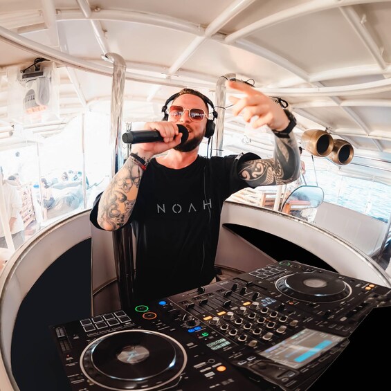 A person with tattoos, wearing a black shirt with 'NOAH' on it, DJing on a boat with a sound system. 5