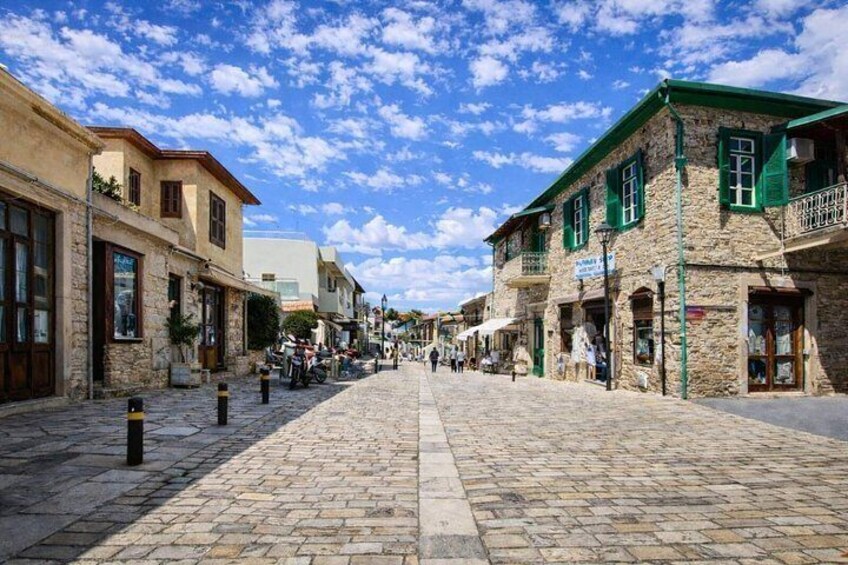 A cobblestone street lined with stone buildings, some with green shutters, under a partly cloudy sky. 1