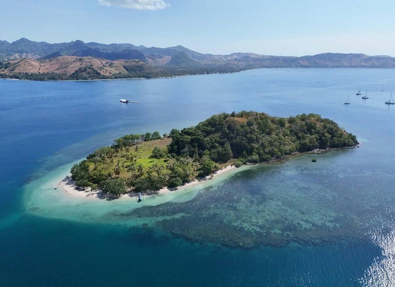 An aerial view of a lush, green island surrounded by clear blue waters with a few boats in the distance. 1