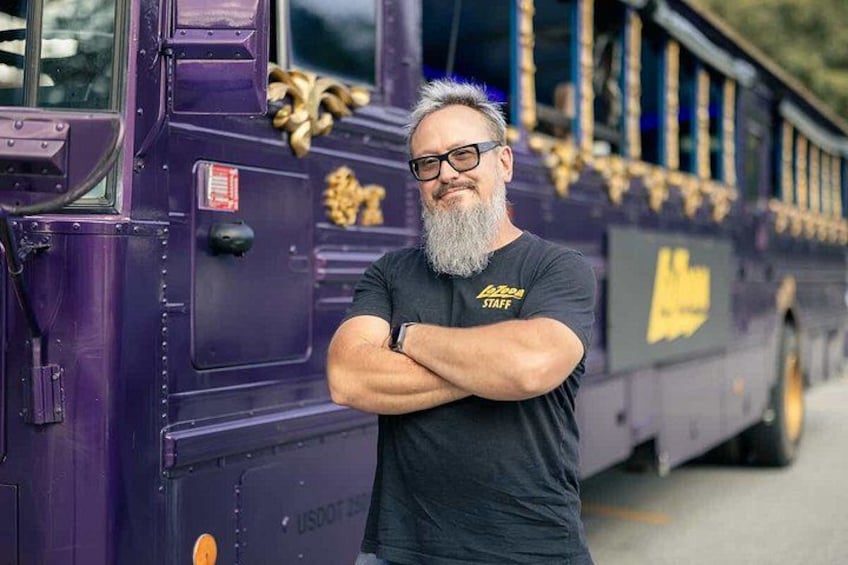 A person standing in front of a purple bus with gold accents and the text 'Staff' on the side. 3
