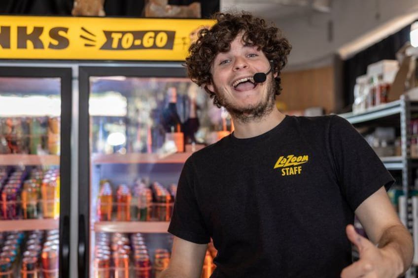 Una persona con camiseta negra que lleva la inscripción 'LoZoom Staff', parada frente a un mostrador refrigerado con diversas bebidas. 3