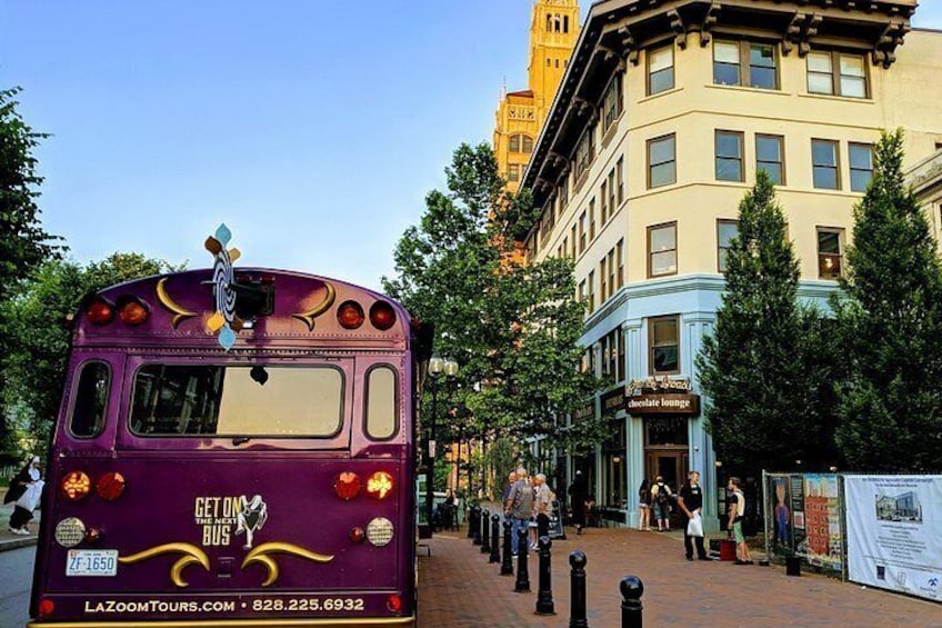 A purple bus with 'Get on the Bus' and 'LAZDOOMTOURS.COM' on the back, parked on a brick sidewalk with a building and trees in the background. 5