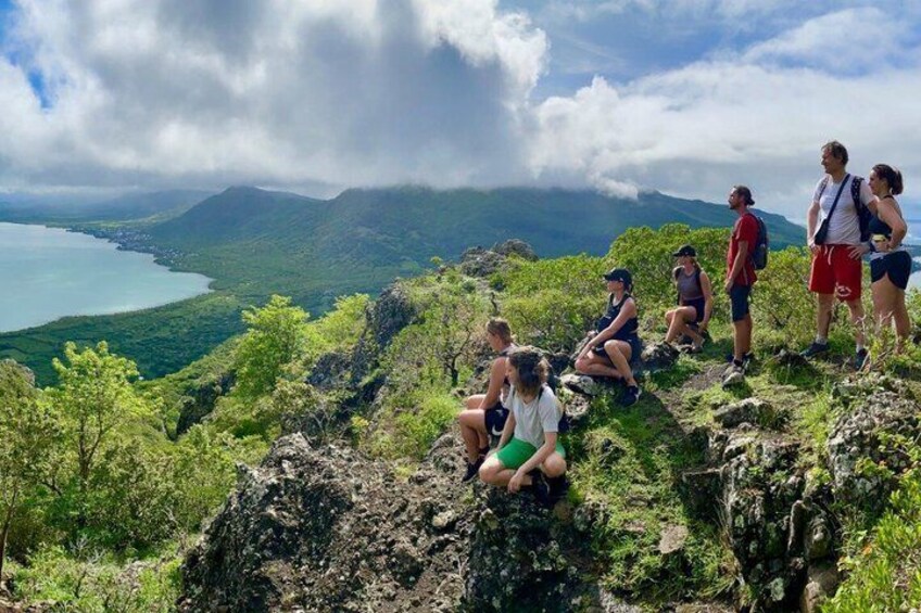 A group of people sitting and standing on a rocky hillside overlooking a coastal landscape with mountains in the distance. 5