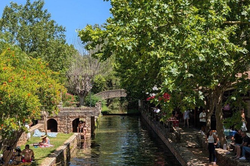 A canal with a stone bridge, trees, and people relaxing by the water. 5