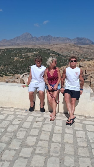 Three people sitting on a stone wall with a mountainous landscape in the background. 3