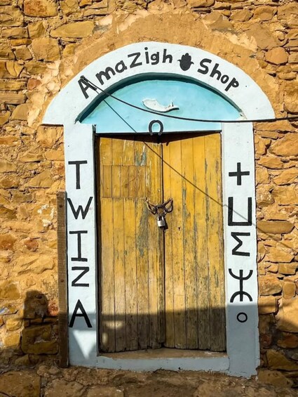 A stone wall with a wooden door labeled 'Amazigh Shop' and 'TWIZA' on either side. 2