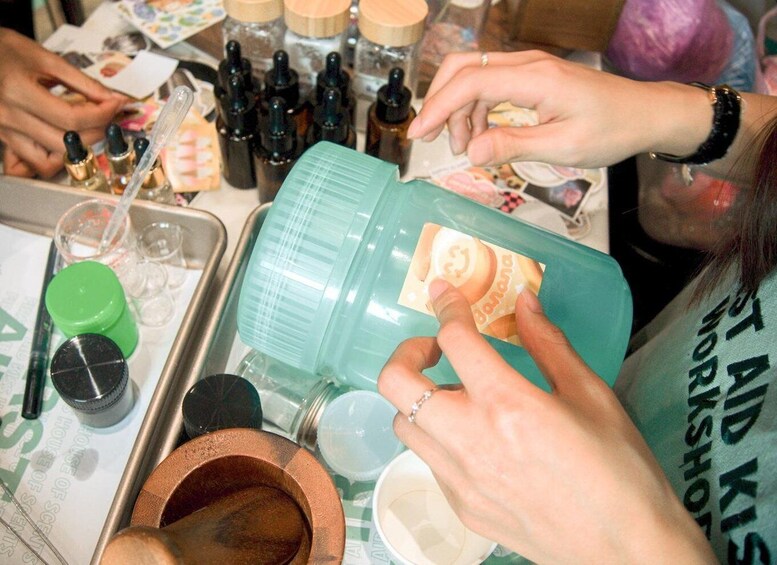 A person holding a green container with a label, surrounded by various bottles and containers on a table. 3