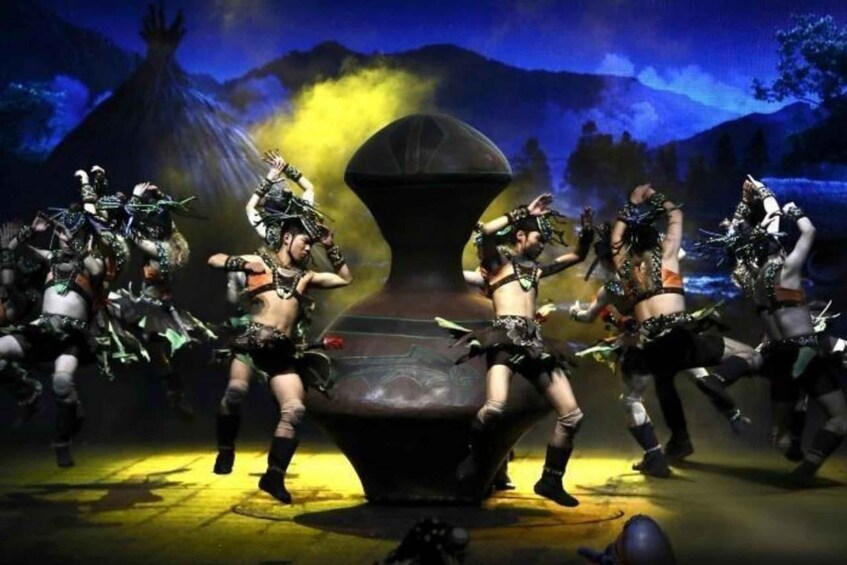 A group of dancers in elaborate costumes performing on a stage with a large vase centerpiece. 2