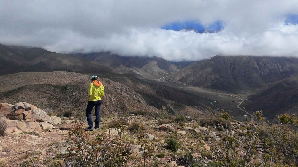 A hiker on a rocky mountain trail with a valley and clouds in the background. 1