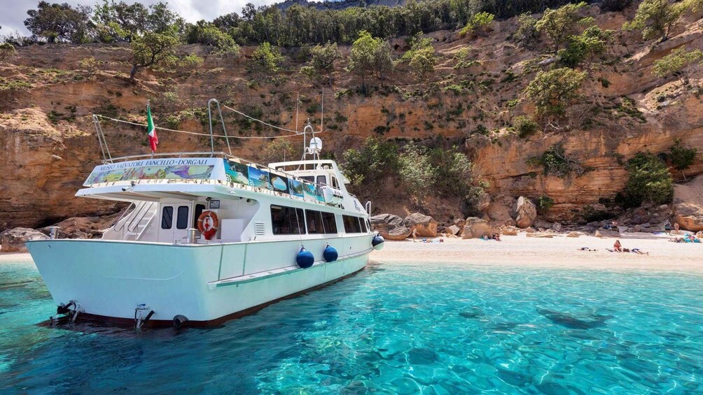 A boat with a lifebuoy and a flag, anchored near a beach with clear turquoise water and rocky cliffs. 4