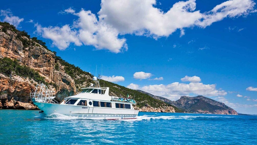 A white boat with the registration number VE8874 on a clear blue sea, with rocky cliffs and mountains in the background. 1