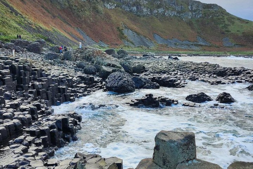 A rocky coastal landscape with waves crashing against large boulders and a steep hillside in the background. 4