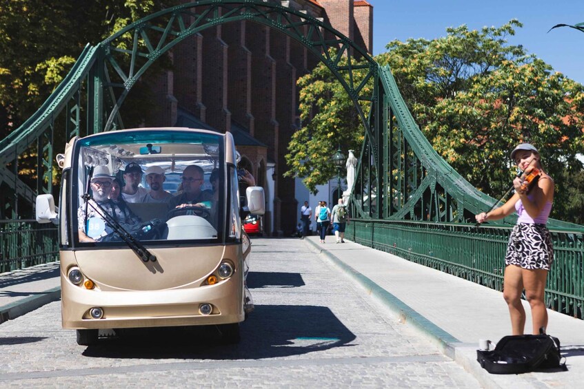 A bus driving on a bridge with a person playing a violin on the sidewalk. 1