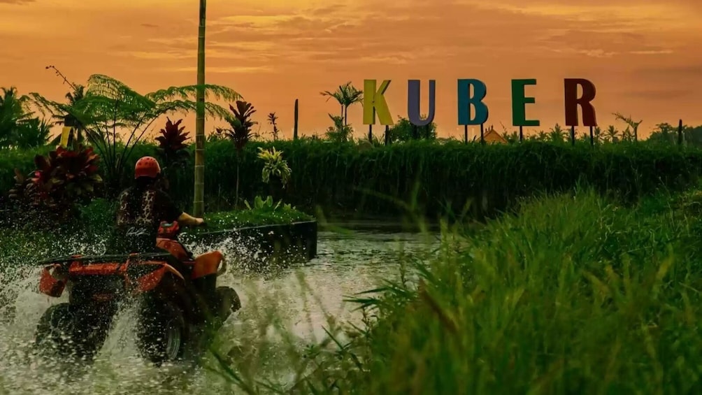 A person riding a red ATV through a water channel with the word 'KUBER' in the background. 1