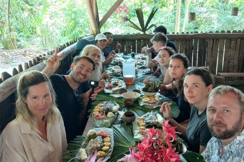 A group of people dining at an outdoor wooden table with various dishes and a tropical setting. 3