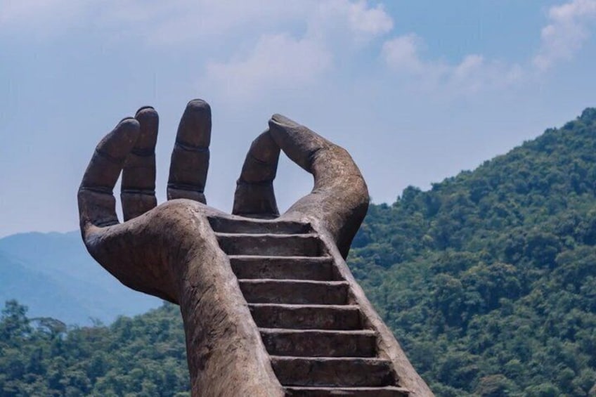 A large hand sculpture with a staircase leading up to it, set against a backdrop of a forested mountain. 4
