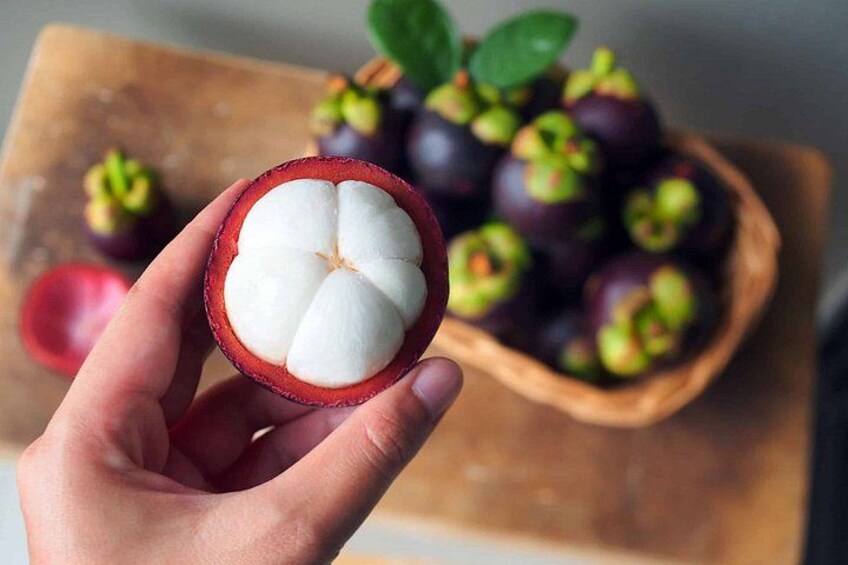A hand holding a half-cut mangosteen fruit with a bowl of whole mangosteens in the background. 5