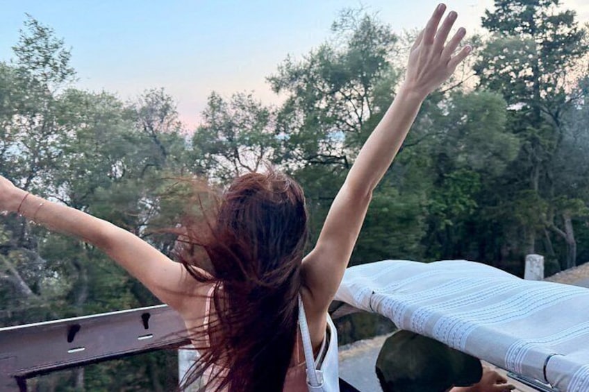 A person with arms raised, wearing a white top, standing on a balcony with a view of trees and a clear sky. 3