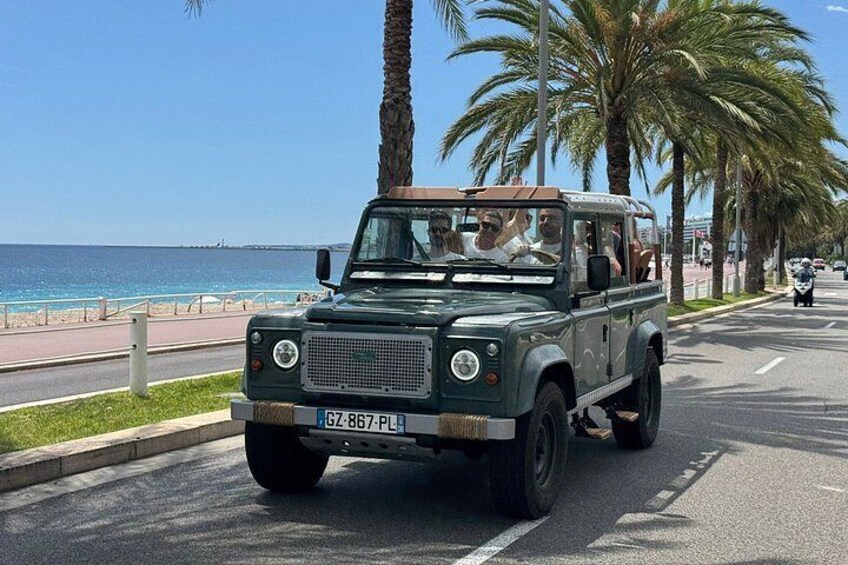 A green off-road vehicle driving on a road with palm trees and a beach in the background. 4