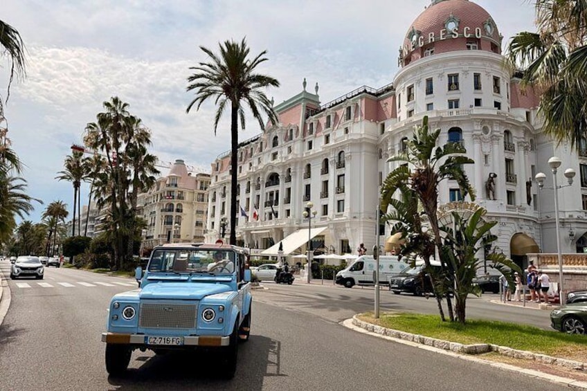 A blue vintage car driving on a street lined with palm trees and a large white building with a dome. 1