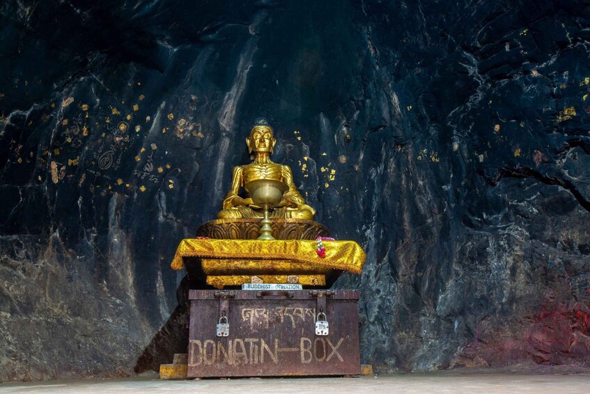En gylden Buddha-statue på en piedestal med inskriptioner, foran en mørk, klippefyldt baggrund. 2