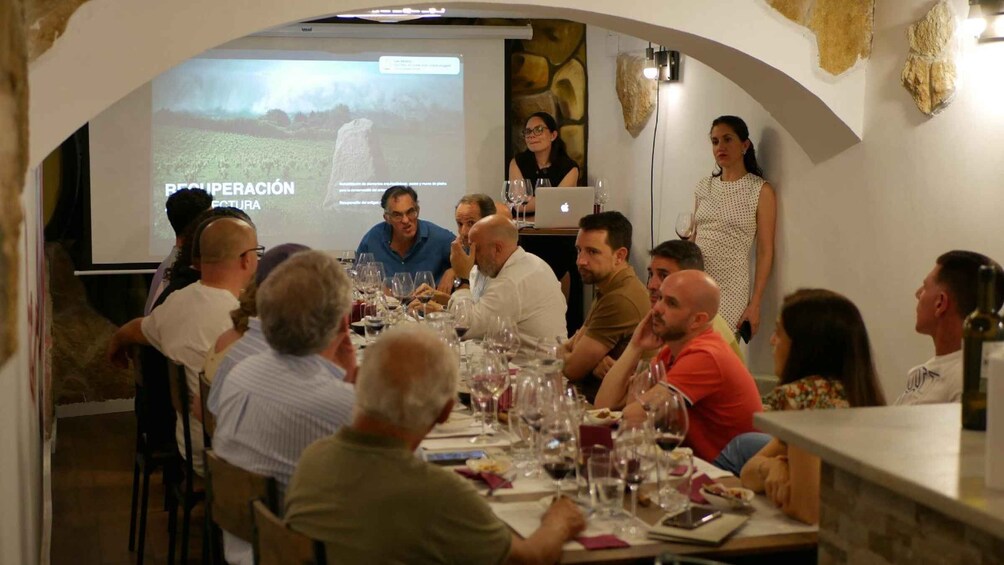 A group of people seated at a table with wine glasses, engaged in a discussion. 1