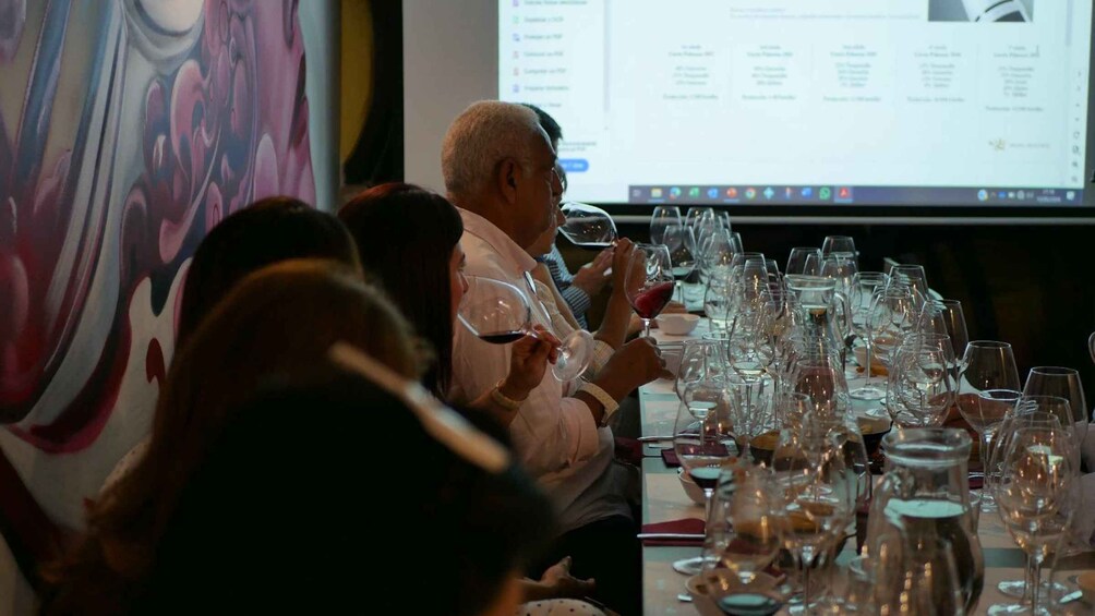 A group of people seated at a table with wine glasses, a projector screen in the background, and a colorful painting on the wall. 4