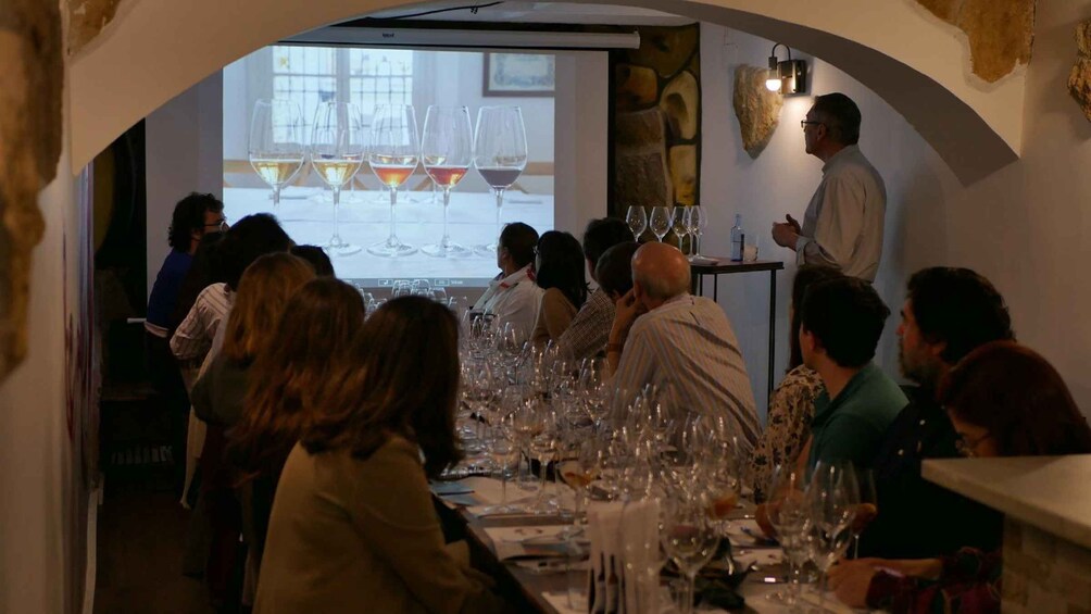 A group of people seated at a long table with wine glasses, in a room with a stone wall and a projector screen displaying wine glasses. 3