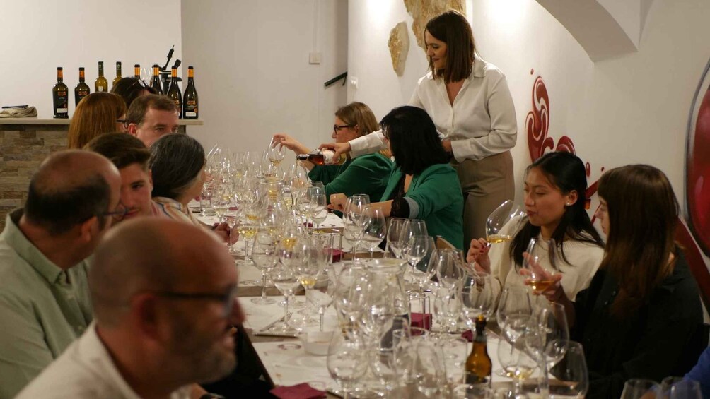 A group of people seated at a long table with multiple wine glasses, some filled with wine, in a restaurant setting. 2