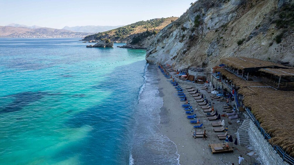 A coastal beach with clear turquoise waters, a row of sun loungers, and a rocky cliff. 2