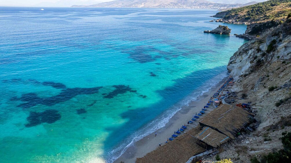 A coastal beach with clear turquoise waters, rocky cliffs, and a row of beach huts. 3