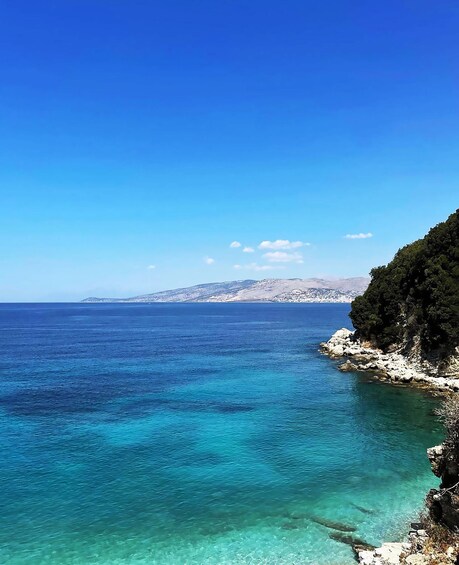 A coastal landscape with clear turquoise waters, rocky cliffs, and a distant mountain range under a clear blue sky. 4