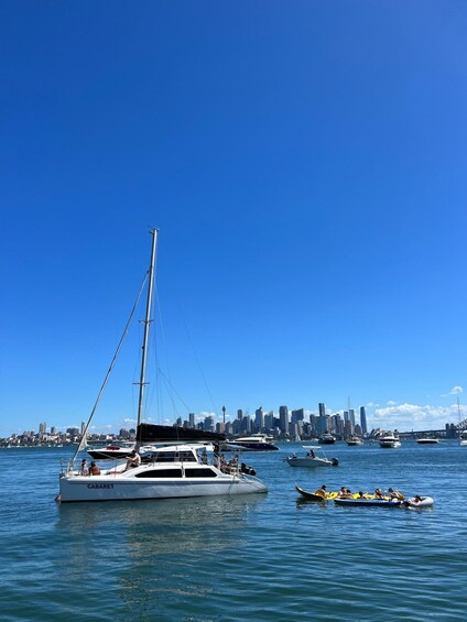 A sailboat named CABARET docked near a city skyline with several other boats and a group of people on a raft in the water. 2