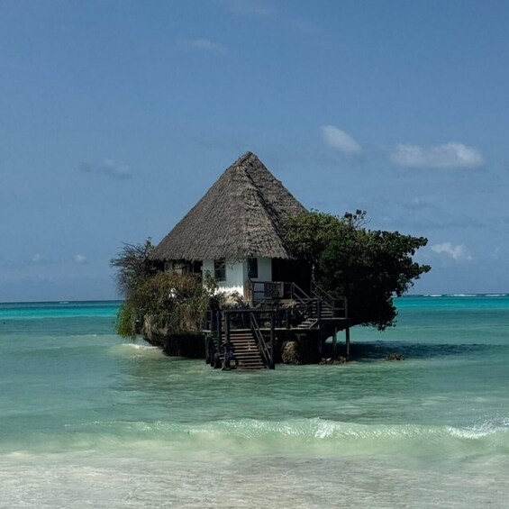 A thatched-roof overwater bungalow with a staircase leading to a deck, surrounded by turquoise water and clear skies. 1