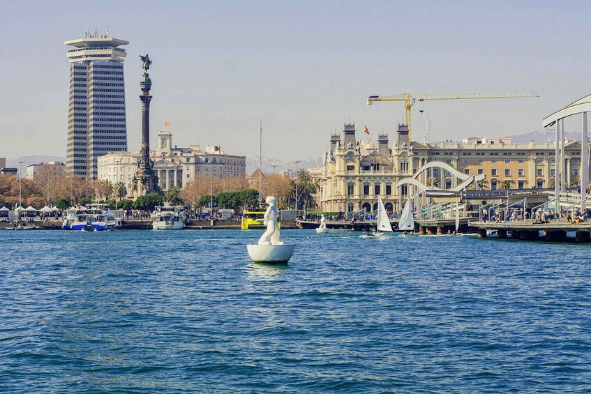 A waterfront with a statue in the water, a tall building, and a bridge with people on it. 2