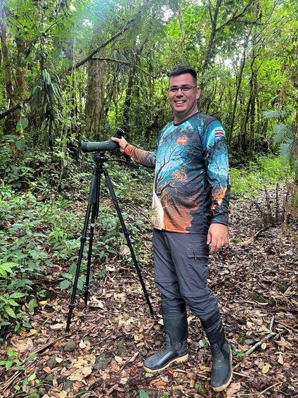 A person standing next to a tripod in a forested area with dense foliage and leaf-covered ground. 4